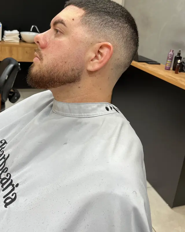 Degradê baixo com barba feita, visto de perfil, com a capa bordada da Barbearia Wolf